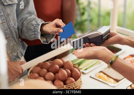 Zugeschnittenes Bild einer vietnamesischen Barista, die das POS-Terminal hält und Kreditkartenzahlungen von Kunden akzeptiert, während sie am Schalter im Café stehen Stockfoto