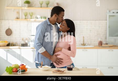 Schwarzer Ehemann berührt den Bauch seiner schwangeren Frau und macht Essen, während er zu Hause in der Küche steht Stockfoto