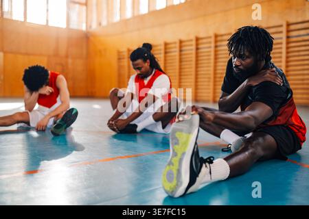 Schwarzer männlicher Basketballspieler, der sich in der Nähe von Teamkollegen aufhält, die auf dem Hallenplatz sitzen Stockfoto