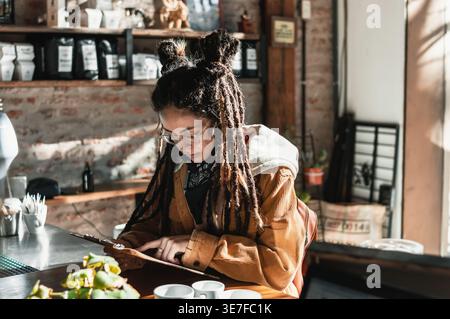 Hispanische Frau mit Dreadlocks, die eine Brille und ein schwarzes Bandana am Hals trug und ein Menü liest, das sich entscheidet, was man essen soll, während man am Tresen im Café steht Stockfoto