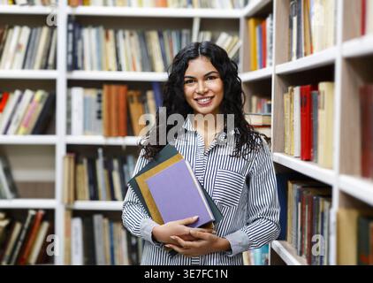 Lächelnde hispanische Studentin, die Bücher trägt, während sie in der Bibliothek steht Stockfoto