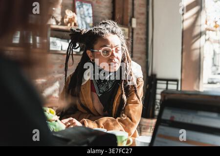Hispanische Frau mit Dreadlocks, Brille und schwarzem Bandana am Hals, die seitlich aussah, während sie an der Theke stand und im Café Essen bestellte Stockfoto