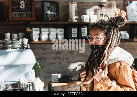 Hispanische Frau mit Dreadlocks mit Brille und schwarzem Bandana am Hals, die an der Theke steht und im Café Essen bestellt Stockfoto