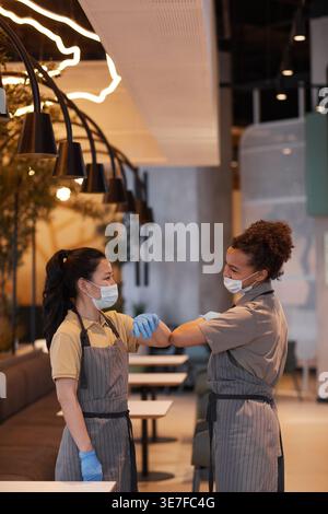 Gemischte und asiatische Kellnerinnen mit Gesichtsmasken und Schürzen, die sich gegenseitig am Ellenbogen stoßen, während sie während der Pandemie im Café stehen Stockfoto