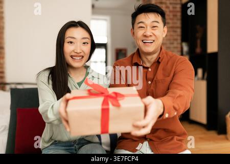 Asiatisches Paar, das die Geschenkbox zusammenhält, während es zu Hause auf dem Sofa sitzt Stockfoto