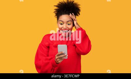 Überraschte schwarze Frau, die einen roten Pullover trägt, indem sie das Smartphone benutzt, um den Kopf zu berühren, während eine Hand vor gelbem Hintergrund im Studio steht Stockfoto