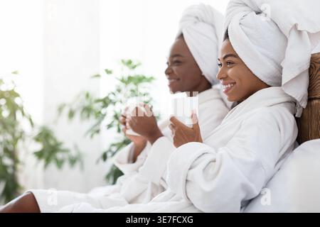 Lächelnde schwarze Frauen mit Handtüchern auf dem Kopf, die Bademäntel mit Tassen tragen, während sie zu Hause auf dem Bett sitzen Stockfoto