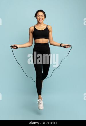 Schwarze Frau in Sportswear, die über das Seil springt, vor blauem Hintergrund im Studio Stockfoto