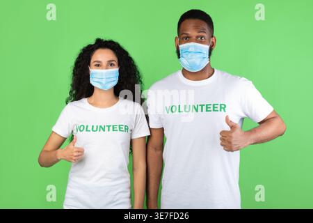 Verschiedene Freiwillige tragen Schutzmasken, die im Studio vor grünem Hintergrund die Daumen nach oben stellen Stockfoto