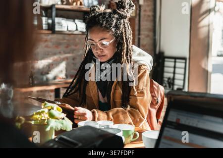 Hispanische Frau mit Dreadlocks, die eine Brille und ein schwarzes Bandana am Hals trug und ein Menü liest, das sich entscheidet, was man essen soll, während man am Tresen im Café steht Stockfoto