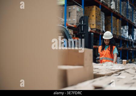 Schwarze Arbeitnehmerin trägt Schutzhelm und Sicherheitsweste, die während der Arbeit im Lager Code auf Kartons scannen Stockfoto