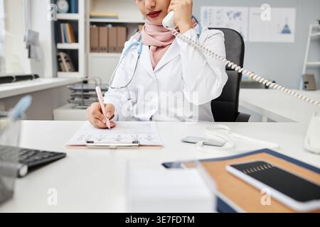 Zugeschnittenes Bild einer Ärztin mit gemischter Rasse in weißem Mantel und Hijab, die einen Stift hält, der über Festnetztelefon telefoniert, während er am Schreibtisch in der Klinik sitzt Stockfoto