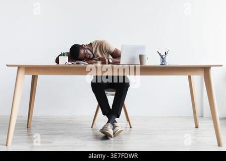Erschöpfter Schwarzer Mann mit Brille, der auf dem Tisch mit Laptop und einer Tasse Kaffee schläft vor der weißen Wand im Home Office Stockfoto