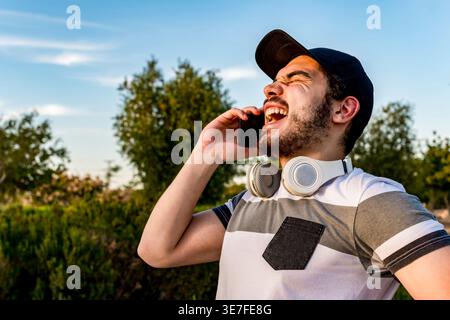 Arabischer Mann mit Mütze und Kopfhörern um den Hals, lachend, während er am Telefon telefoniert, während er am sonnigen Tag im Park steht Stockfoto