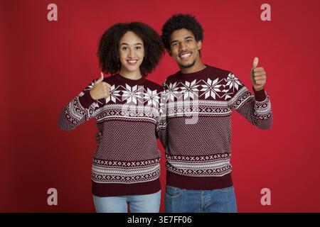 Ein lächelndes schwarzes Paar mit einem roten Pullover, der die Daumen nach oben drückt, während er im Studio vor rotem Hintergrund steht Stockfoto
