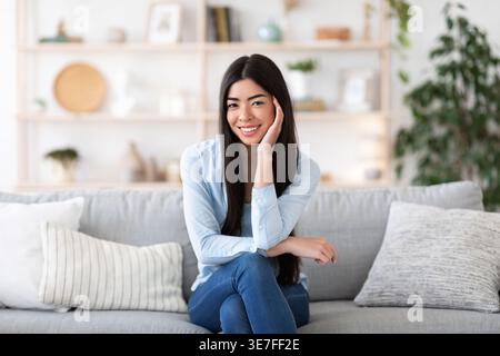 Lächelnde asiatische Frau, die das Kinn auf der Hand anlehnt, während sie mit überkreuzten Beinen im Wohnzimmer zu Hause auf dem Sofa sitzt Stockfoto