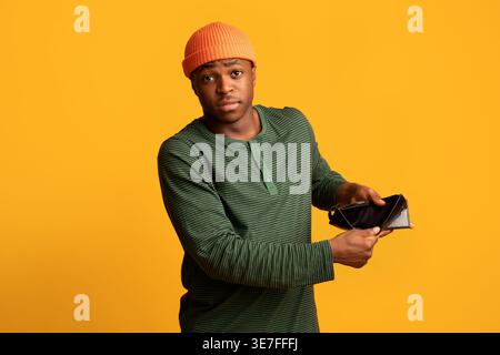 Schwarzer Mann in Beanie, der leere Brieftasche zeigt, während er im Studio vor gelbem Hintergrund steht Stockfoto