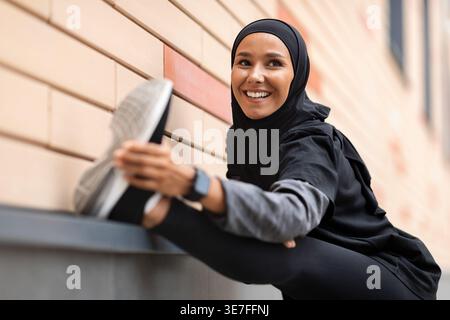 Lächelnde muslimische Frau aus dem Nahen Osten, die Hijab und Sportkleidung trägt und die Beine ausstreckt, während sie vor einer Ziegelmauer auf der Straße steht... Stockfoto