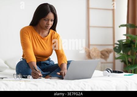 Serious Black Studentin schreibt in Notizbuch studiert und benutzt Laptop mit überkreuzten Beinen auf dem Bett zu Hause Stockfoto