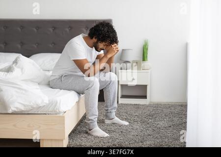 Gestresster bärtiger indischer Mann mit lockigem Haar, der die Stirn auf geballten Fäusten lehnt und im Schlafzimmer zu Hause auf dem Bett sitzt Stockfoto