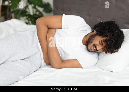 Kranker asiatischer Mann mit lockigen Haaren, der den Bauch berührt und unter Schmerzen leidet, während er mit geschlossenen Augen auf dem Bett zu Hause liegt Stockfoto