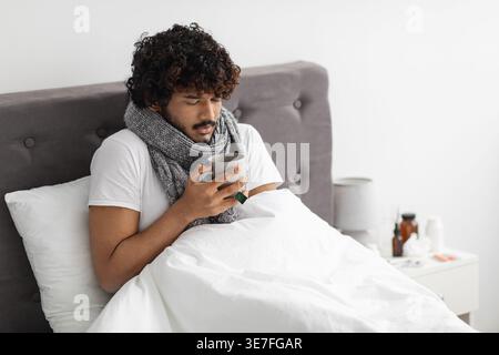 Ein kranker asiatischer Mann mit lockigen Haaren, der einen Schal um den Hals trägt, der eine Tasse Kaffee hält, während er zu Hause im Bett sitzt Stockfoto