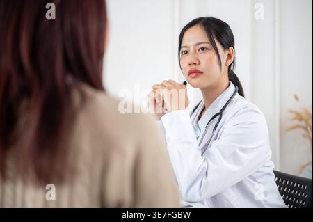 Thailändische Ärztin mit gefesselten Händen, die ein Stethoskop am Hals trägt und den Stift hält, hört der weiblichen Patientin zu, die ihre Symptome am Tisch ... erklärt Stockfoto