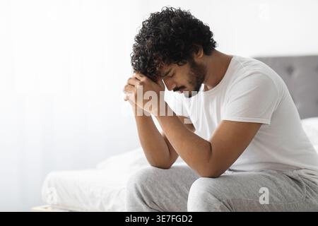 Gestresster bärtiger indischer Mann mit lockigem Haar, der die Stirn auf geballten Fäusten lehnt und im Schlafzimmer zu Hause auf dem Bett sitzt Stockfoto