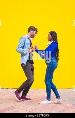 Lächelnde Schwarze und kaukasische Paare mit Kopfhörern um den Hals halten Hände zusammen tanzen vor gelbem Hintergrund im Studio Stockfoto