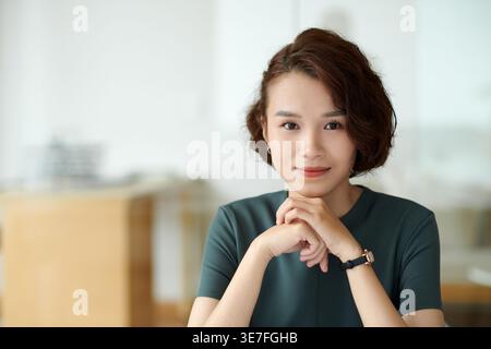 Asiatische Geschäftsfrau, die das Kinn auf gefaltete Hände stützt und am Tisch im Büro sitzt Stockfoto