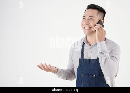 Lächelnder asiatischer männlicher Barista mit Schürze und Gesten, der auf dem Smartphone spricht und im Studio vor weißem Hintergrund steht Stockfoto