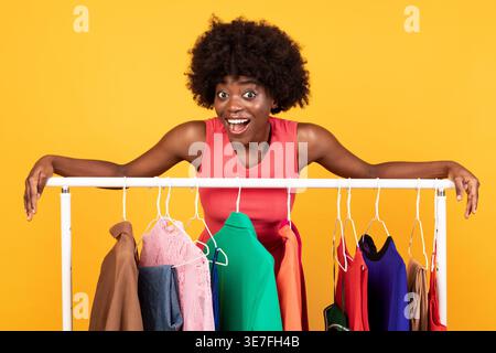 Schwarze Frau mit Afro und offenem Mund, die sich auf einen Kleiderständer stützt, während sie im Studio vor gelbem Hintergrund steht Stockfoto