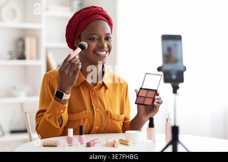 Schwarze Frau mit Kopftuch, die am Tisch sitzt und ein Video aufnimmt, wie sie einen Pinsel hält und zu Hause Make-up aufträgt Stockfoto