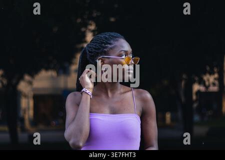 Schwarze Frau mit Zöpfen, die eine Sonnenbrille und ein lila Kleid tragen und tagsüber seitlich auf der Straße stehen Stockfoto