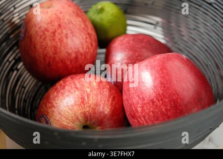 Eine Nahaufnahme mehrerer reifer roter Äpfel und einer einzelnen grünen Limette, die in einer modernen schwarzen dekorativen Obstschale aus Draht ruhen. Stockfoto