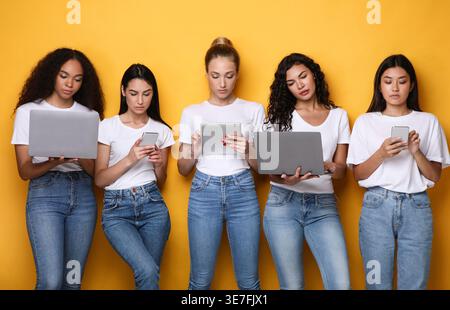 Verschiedene Frauengruppen, die Hemden und Jeans mit verschiedenen elektronischen Geräten tragen und im Studio vor gelbem Hintergrund stehen Stockfoto