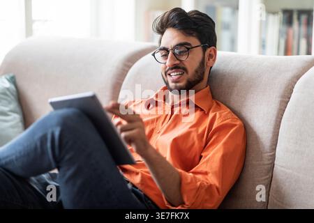 Entspannter cooler, stilvoller junger Mann aus dem Nahen Osten, der im Wohnzimmer auf der Couch sitzt, zu Hause ein digitales Tablet verwendet und lächelt, mit Freunden chatet, Websur Stockfoto