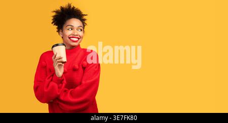 Schwarze Frau, die einen roten Pullover trägt, der eine Tasse Kaffee hält, mit einer Hand, die vor gelbem Hintergrund im Studio steht Stockfoto