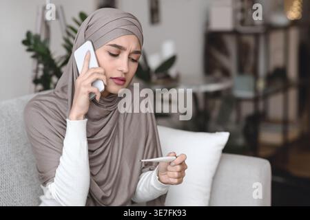 Kranke arabische Frau im Hijab, die ein Thermometer hält und den Arzt mit einem Smartphone anruft, das zu Hause auf dem Sofa sitzt Stockfoto
