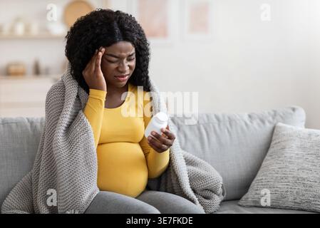 Kranke Schwarze schwangere Frau mit lockigen Haaren, die gelbe lange Ärmel trägt, die in eine Decke gewickelt sind, die den Tempel berührt und auf der Arzneimittelflasche sitzt... Stockfoto