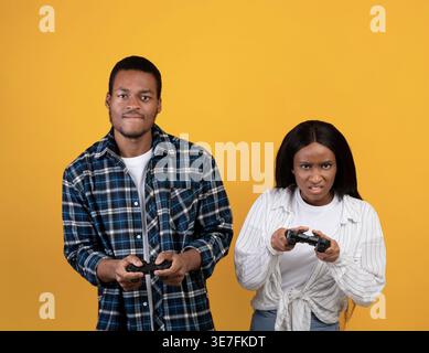 Schwarzes Paar, das Spielecontroller hält, während es im Studio vor gelbem Hintergrund steht Stockfoto