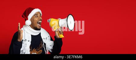 Schwarzer Mann mit Weihnachtsmann-Hut und Pullover, der wegschaut und auf Megaphon brüllt, steht mit offener Hand vor rotem Hintergrund im Studio Stockfoto