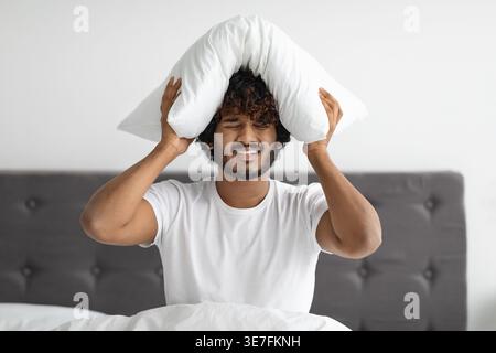 Kranker asiatischer Mann mit lockigen Haaren, der seinen Kopf mit Kissen bedeckt, während er zu Hause auf dem Bett sitzt Stockfoto