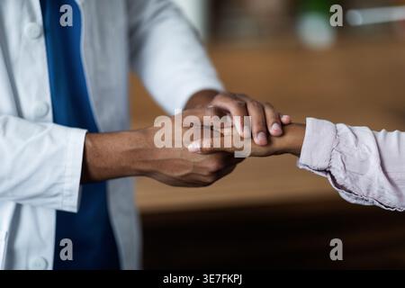 Zugeschnittenes Bild eines schwarzen männlichen Arztes, der die Hände des Patienten hält, der vor dem Tisch in der Klinik steht Stockfoto