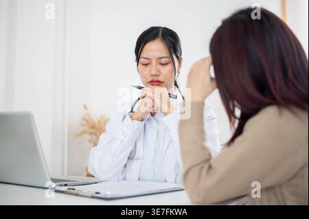 Thailändische Ärztin mit gefesselten Händen am Kinn hält Stift nach unten und hört weiblichen Patienten zu, die ihre Symptome am Tisch mit la... Stockfoto