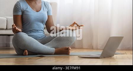 Schwarze Frau, die Online-Meditations-Yoga-Kurs hat, sitzt in Lotusposition vor Laptop im Wohnzimmer zu Hause Stockfoto