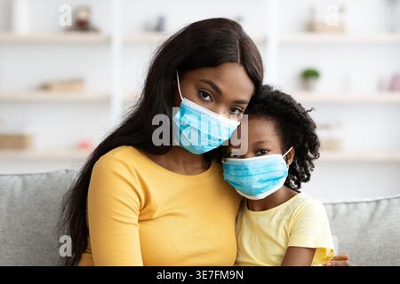 Schwarze Mutter und Tochter tragen Gesichtsmasken und sitzen dicht zusammen auf dem Sofa im Wohnzimmer zu Hause Stockfoto