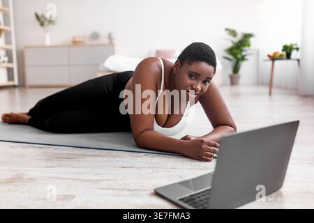 Erschöpfte kurvige schwarze Frau, die auf Yogamatte in der Nähe des Laptops im Schlafzimmer zu Hause liegt Stockfoto