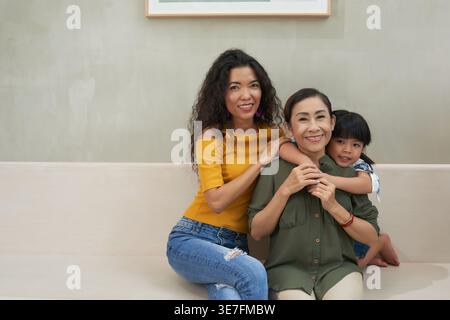 Asiatische Großmutter wird von Tochter und Enkelin umarmt, die zu Hause zusammen auf dem Sofa sitzen Stockfoto