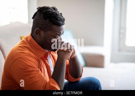 Ernster Schwarzer Mann, der den Mund auf gefesselte Hände lehnt, sitzt auf Sofa im Wohnzimmer zu Hause Stockfoto